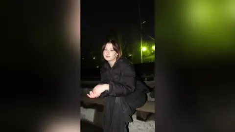 Young woman in a black puffer jacket sitting outdoors at night under streetlights with a moody atmosphere.