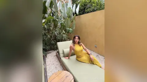 Young woman in a yellow summer dress relaxing on a lounge chair in a sunny tropical garden patio.