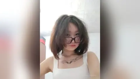 TishaOyervides wears thick black glasses and a white tank top, showing her dark hair and chest tattoo against a plain white wall.