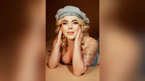 FiorellaRossi lies on her stomach wearing a light blue beret and denim shorts, resting her chin on her hands while looking at the camera.
