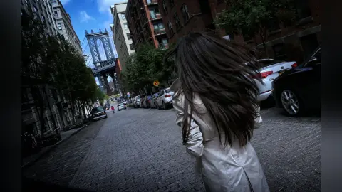 Woman in a white trench coat walking down a city street near the Manhattan Bridge on a breezy day.