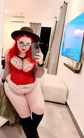 Redheaded woman in a red bodysuit and fishnet sleeves taking a mirror selfie in a modern living room.