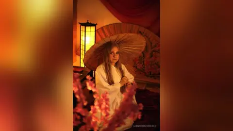 Young woman in a white flowing dress holding a straw hat in a warm, moody indoor setting with soft lighting.