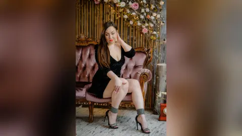 TannySmith sits on a pink velvet sofa wearing a short black dress and high heels, posing with her hand in her long dark hair.