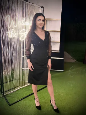 Elegant woman in a black long-sleeve cocktail dress posing by a glowing neon sign on a green lawn at night.