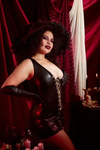 Glamorous woman in a black latex corset dress and long gloves posing against red velvet curtains.