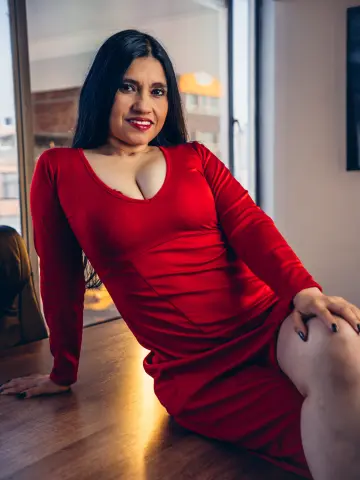 Smiling woman in a red long-sleeved dress sitting on a wooden floor in a bright modern room.