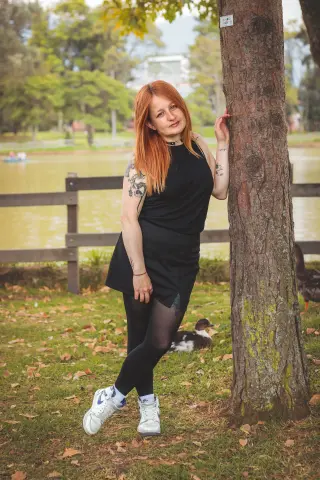 Red-haired woman in a black dress leaning against a tree by a lake in a peaceful park setting.