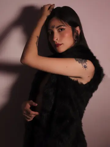 Tattooed woman in a black faux fur vest posing with moody lighting against a pink background.