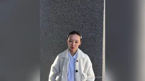 Young woman in a cozy white fleece jacket posing against a textured grey wall in soft natural light.