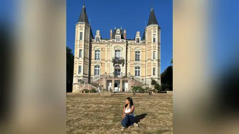 Young woman in a white top and jeans sitting on a sunny lawn in front of a grand historic French chateau.