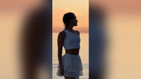 Silhouette of a woman in a white summer outfit standing by the ocean during a peaceful golden sunset.