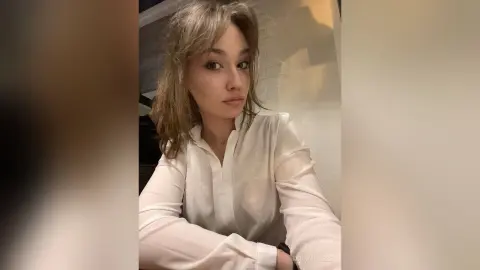 Young woman with wavy blonde hair wearing a white button-down shirt in a soft, moody indoor setting.