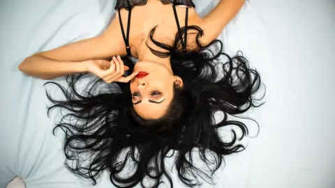 OliviaLarson lies on a white bed with her long black hair spread out, wearing a black lace bra and red lipstick.