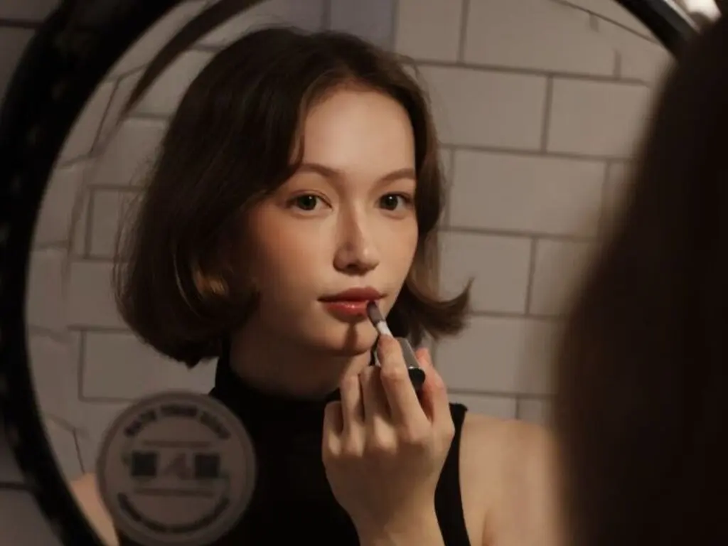 Young woman with short bob hair applying lip gloss in a mirror within a modern bathroom setting.