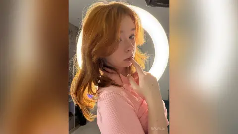 Redheaded woman in a pink ribbed shirt posing under a bright ring light for a soft, glowing portrait.