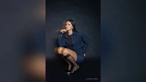 Thoughtful woman in a navy striped oversized shirt and black shorts posing against a dark studio background.