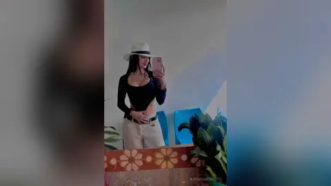 Stylish woman in a black long-sleeve top and white fedora taking a mirror selfie in a bright room.