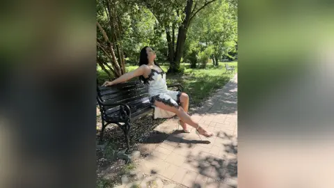 Elegant woman in a white lace dress sitting on a park bench under sunny green trees.