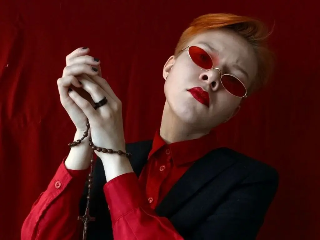 Redheaded woman in a red shirt and black blazer posing with hands clasped against a red backdrop.