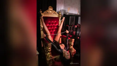 Woman in black leather outfit sitting on a gold ornate throne holding chains in a moody, dark room.