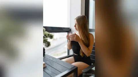 Relaxed woman in black bodysuit sitting by a bright window holding a coffee mug in a peaceful morning setting.