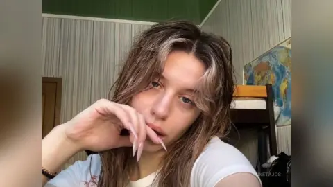 Young woman with wavy brown hair in a white t-shirt posing in a cozy bedroom setting.