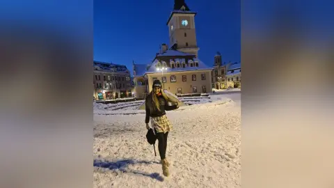 TiffanyAddams stands in a snowy outdoor setting at night, wearing a black top and a plaid skirt or kilt.