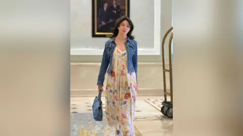 MnemoZeina wears a long floral print dress and a blue denim jacket while standing in a room with a framed picture and a gold cart.