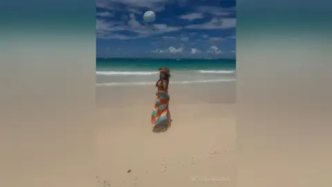 Woman in a colorful maxi dress and sun hat walking on a sunny tropical beach under a blue sky.