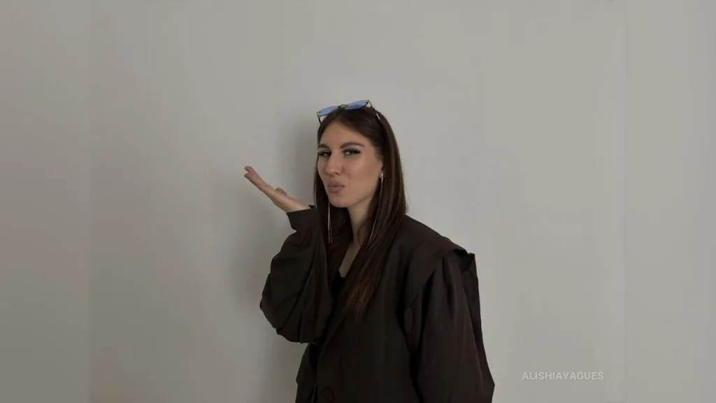 AlishiaYagues poses with her hand touching her cheek, wearing a dark blazer and her hair in half-up buns.
