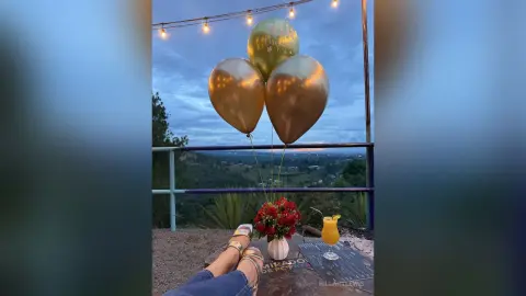 Gold balloons and a cocktail on a balcony overlooking a scenic landscape at sunset.