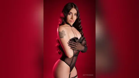 Glamorous woman in black sheer bodysuit and lace gloves posing against a moody red studio background.
