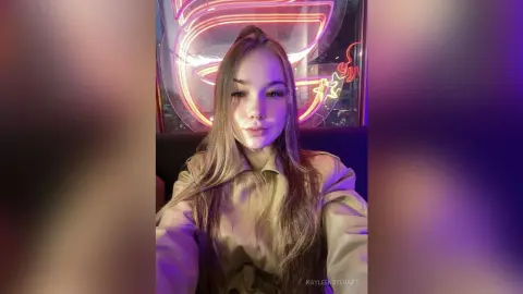 Young woman in a beige turtleneck sweater posing under glowing pink neon lights in a moody setting.