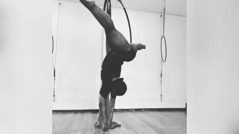 KeianHoz hangs upside down from an aerial hoop, showing her bare legs and athletic body in a black sports bra and shorts.