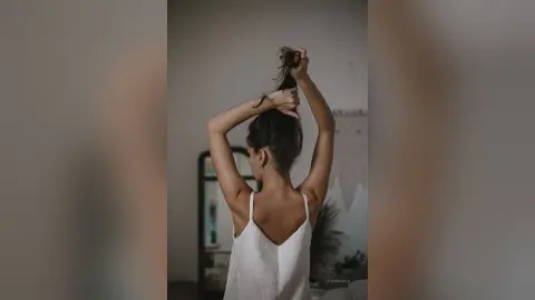 Young woman in a white tank top styling her hair in a minimalist, moody indoor setting.