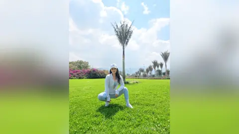 Young woman in light blue athletic wear squatting on a sunny green lawn near palm trees.