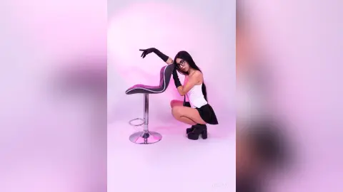 Stylish woman in white tank top and black gloves posing on a stool against a pink studio background.