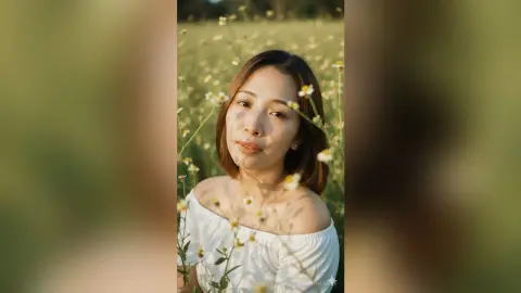 Smiling woman in a white off-the-shoulder top relaxing in a sunny wildflower meadow during golden hour.