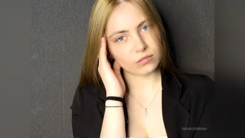 Blonde woman in a black blazer and silver necklace posing thoughtfully against a grey wall.