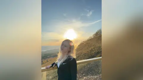 Blonde woman in a black zip-up jacket standing on a scenic coastal overlook during a golden sunset.