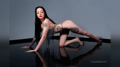 Tattooed woman in a shimmering silver bodysuit posing on a reflective floor in a moody studio setting.