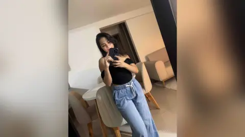 Smiling young woman in a black crop top and blue denim trousers taking a selfie in a bright modern living room.