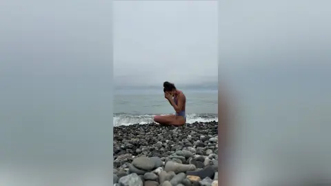 Melancholic woman in a swimsuit sitting on a rocky beach under a cloudy sky.