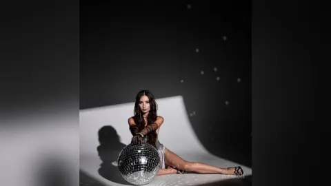 Elegant woman in a silver sequin dress sitting by a disco ball in a moody, minimalist studio setting.