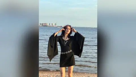 LorenLoiss stands on a sandy beach wearing a short black dress with long flared sleeves and a silver chain belt.
