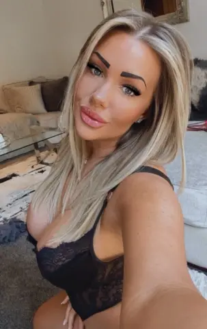Blonde woman in black lace lingerie posing in a cozy living room setting.