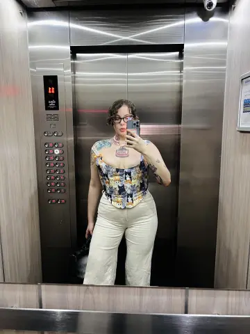 Woman in floral corset top and beige trousers taking a mirror selfie in a modern elevator.