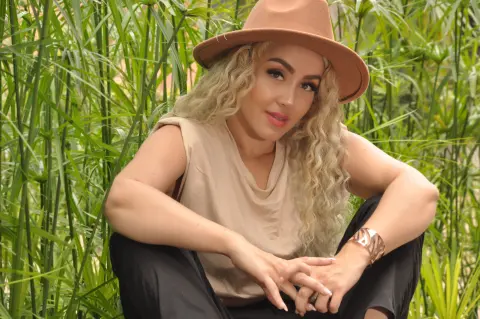 Blonde woman in a tan sleeveless top and brown hat relaxing in a lush green garden setting.