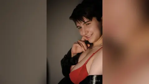 Smiling young woman with short dark hair wearing a red lace bra and black jacket in a moody indoor setting.